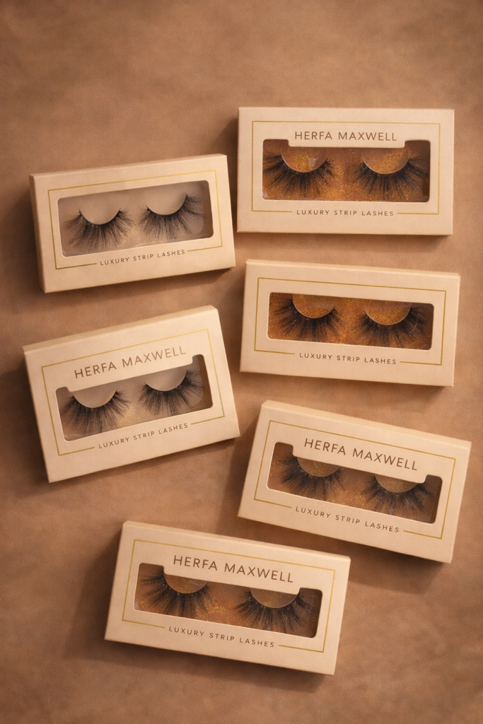 Product flat lay featuring multiple Herfa Maxwell luxury strip lashes in elegant cream packaging with gold accents, arranged on a warm neutral background to showcase different lash styles.
